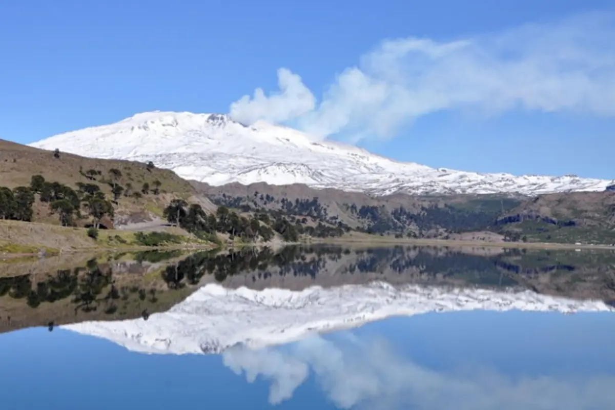 Volcán nevado humeante reflejado en un lago tranquilo rodeado de colinas y vegetación