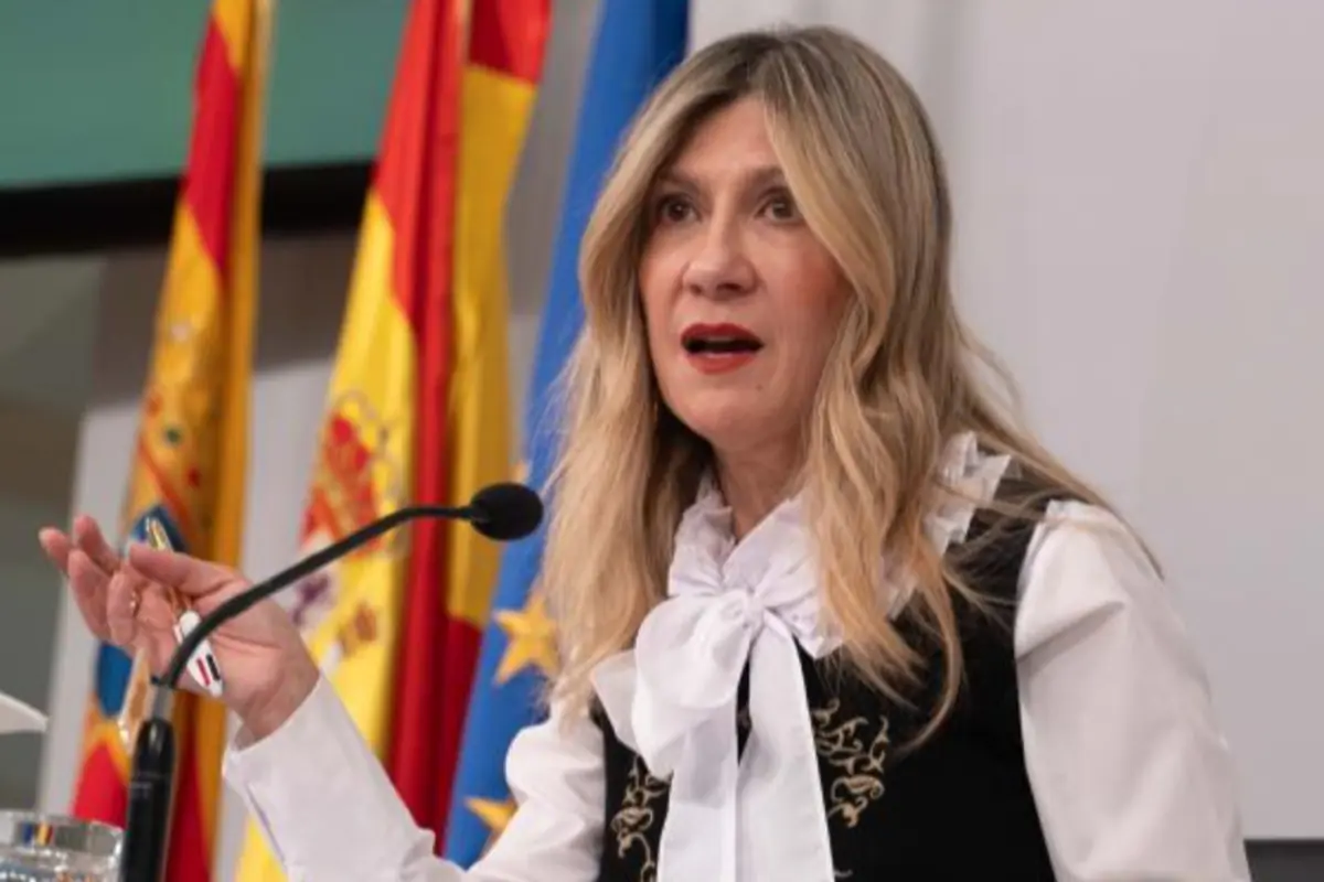 Mujer rubia hablando en una rueda de prensa frente a micrófono con banderas de España y de la Unión Europea al fondo