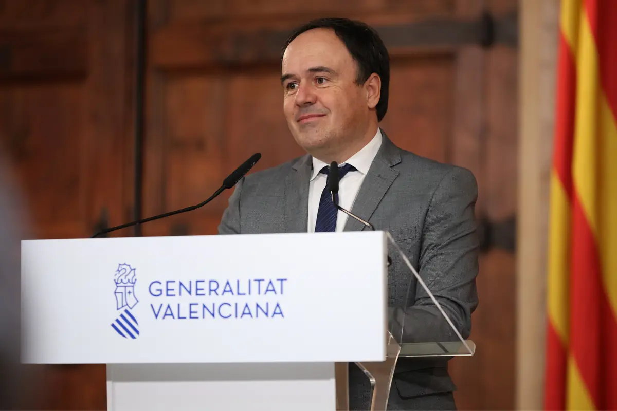 Hombre con traje gris hablando en un atril de la Generalitat Valenciana con micrófonos delante y bandera valenciana al fondo