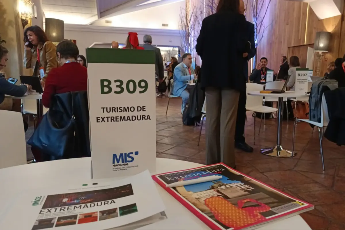 Mesa informativa de Turismo de Extremadura en una feria profesional con folletos sobre la región y varias personas conversando en un espacio de reuniones Mesa informativa de Turismo de Extremadura en una feria profesional con folletos sobre la región y varias personas conversando en un espacio de reuniones