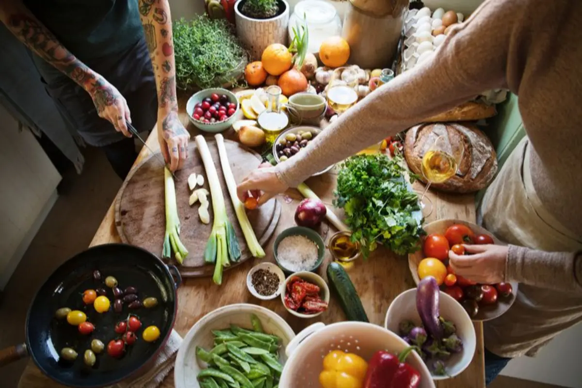 Dos personas preparando una comida con muchas verduras frescas y hierbas sobre una mesa de cocina de madera