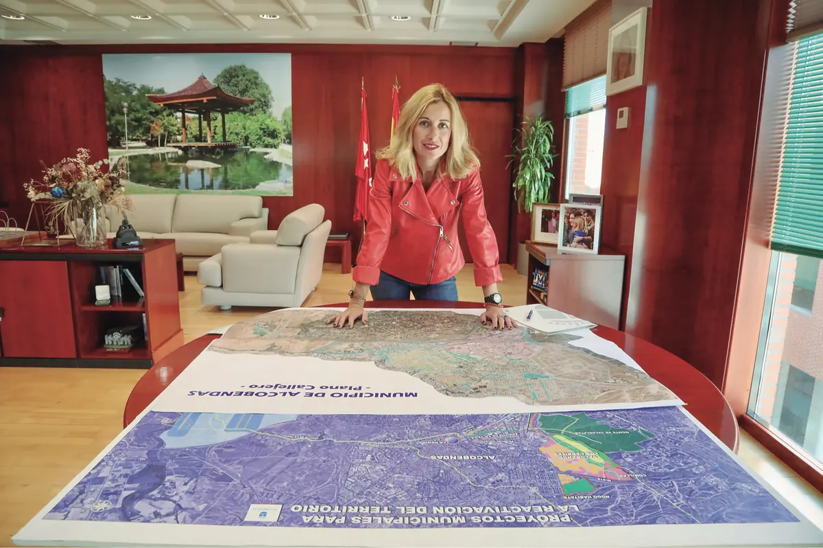 Mujer rubia con chaqueta roja apoyada sobre grandes planos urbanos extendidos en una mesa dentro de una oficina amplia y luminosa con muebles de madera y sofás claros