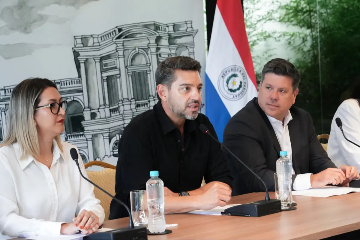 Tres personas sentadas en una mesa con micrófonos durante una conferencia de prensa frente a una bandera de Paraguay