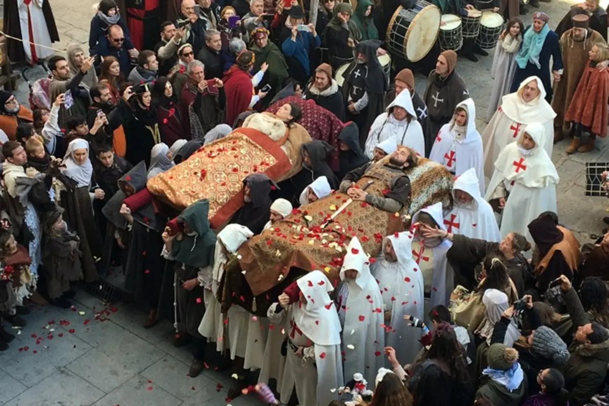 Procesión medieval con dos figuras yacentes sobre andas adornadas con telas bordadas, portadas por personas con túnicas y capuchas mientras una multitud las rodea y lanza pétalos de flores