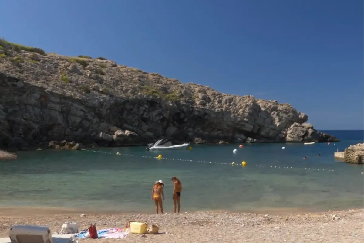 Cala de agua turquesa rodeada de rocas con dos personas en la orilla y una lancha anclada en el mar