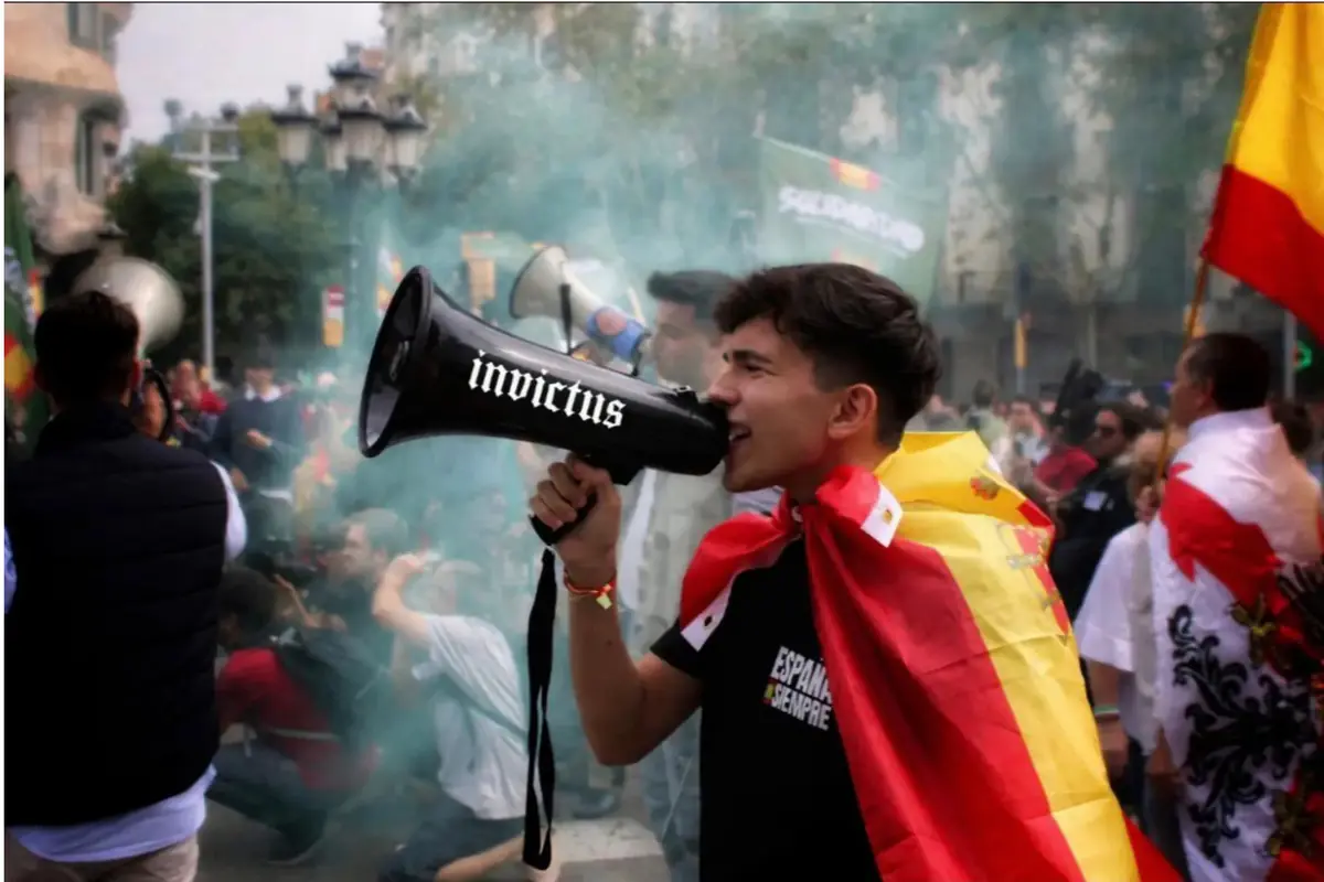 Joven envuelto en una bandera de España gritando por un megáfono con la palabra invictus en medio de una manifestación con humo de colores y más personas alrededor