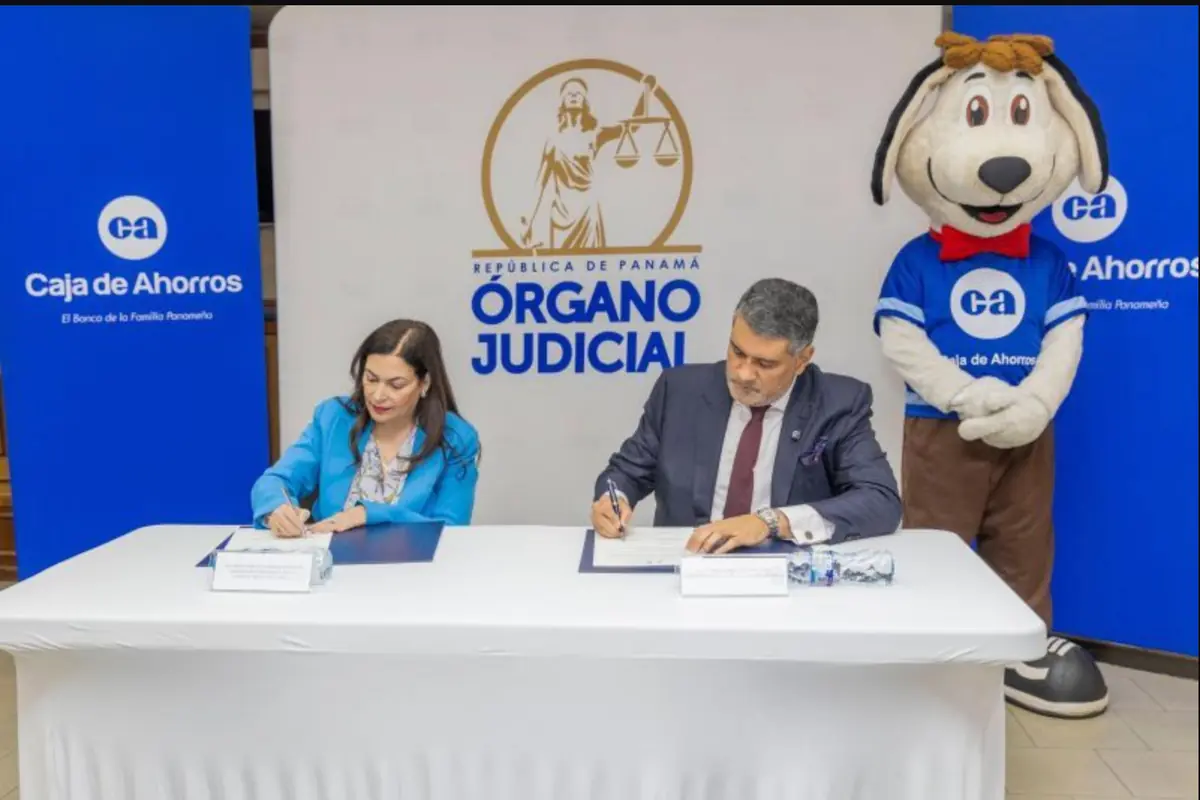 Dos representantes de la Caja de Ahorros y del Órgano Judicial de Panamá firman un convenio en una mesa blanca mientras una mascota con disfraz de perro de la Caja de Ahorros posa a un lado