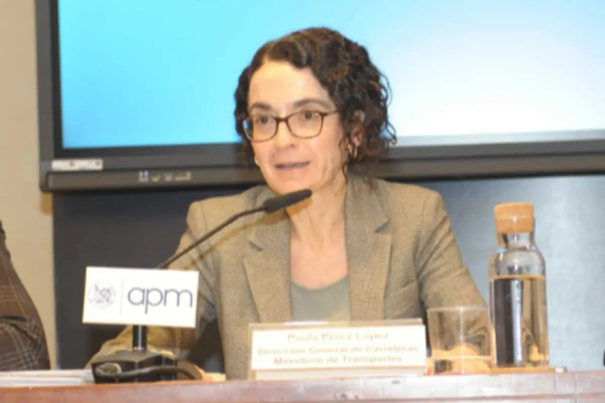 Mujer con gafas y chaqueta gris hablando en una conferencia sentada tras una mesa con micrófono cartel identificativo y botella de agua