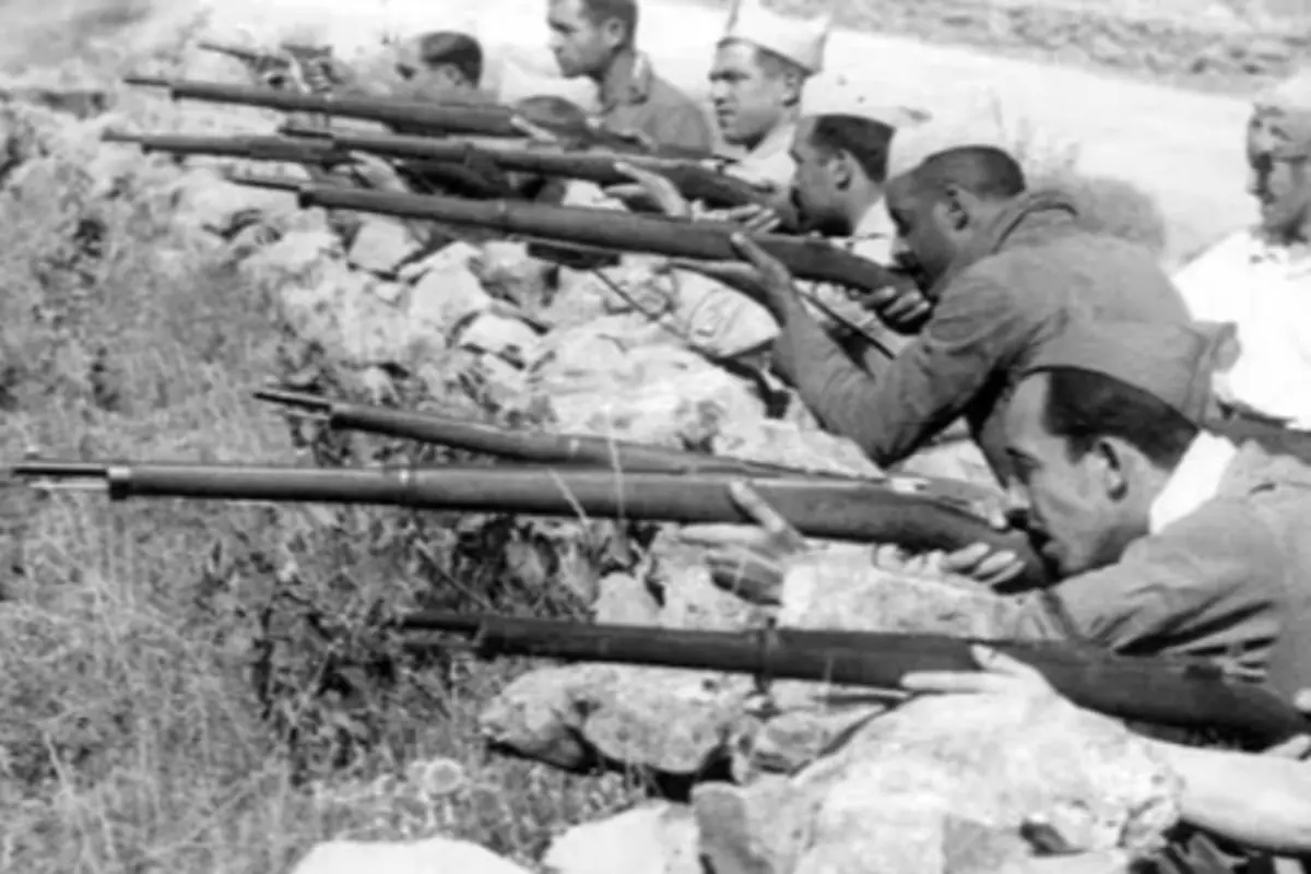 Soldados en blanco y negro apuntan con fusiles desde una trinchera de piedras durante un conflicto armado