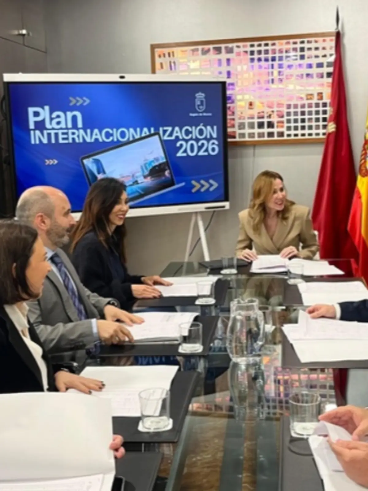 Reunión de trabajo en una sala de conferencias con varias personas sentadas alrededor de una mesa de cristal mientras una pantalla muestra el texto Plan Internacionalización 2026 de la Región de Murcia