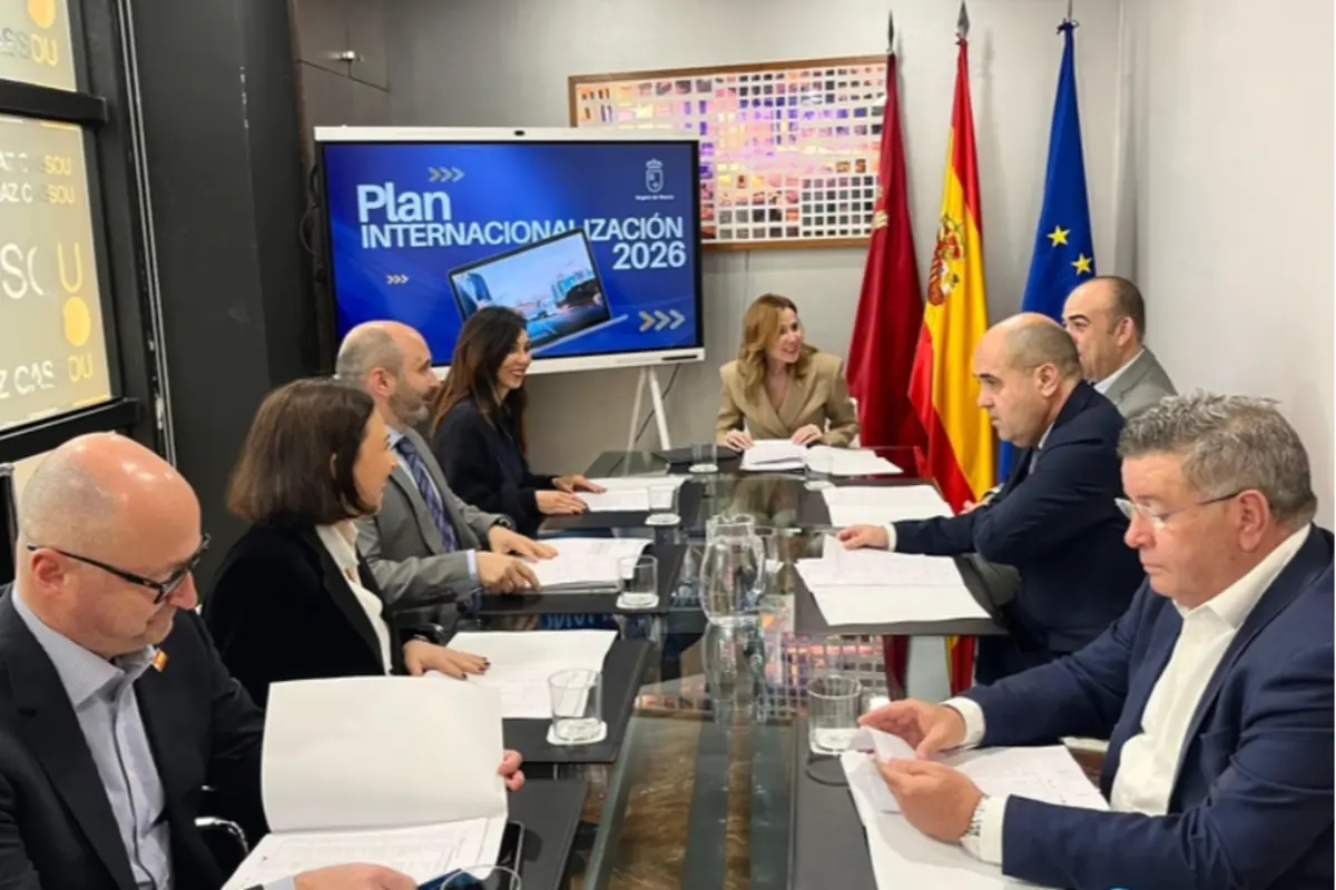 Reunión de trabajo con varias personas sentadas alrededor de una mesa de cristal revisando documentos frente a una pantalla que muestra el texto Plan Internacionalización 2026 y varias banderas oficiales al fondo