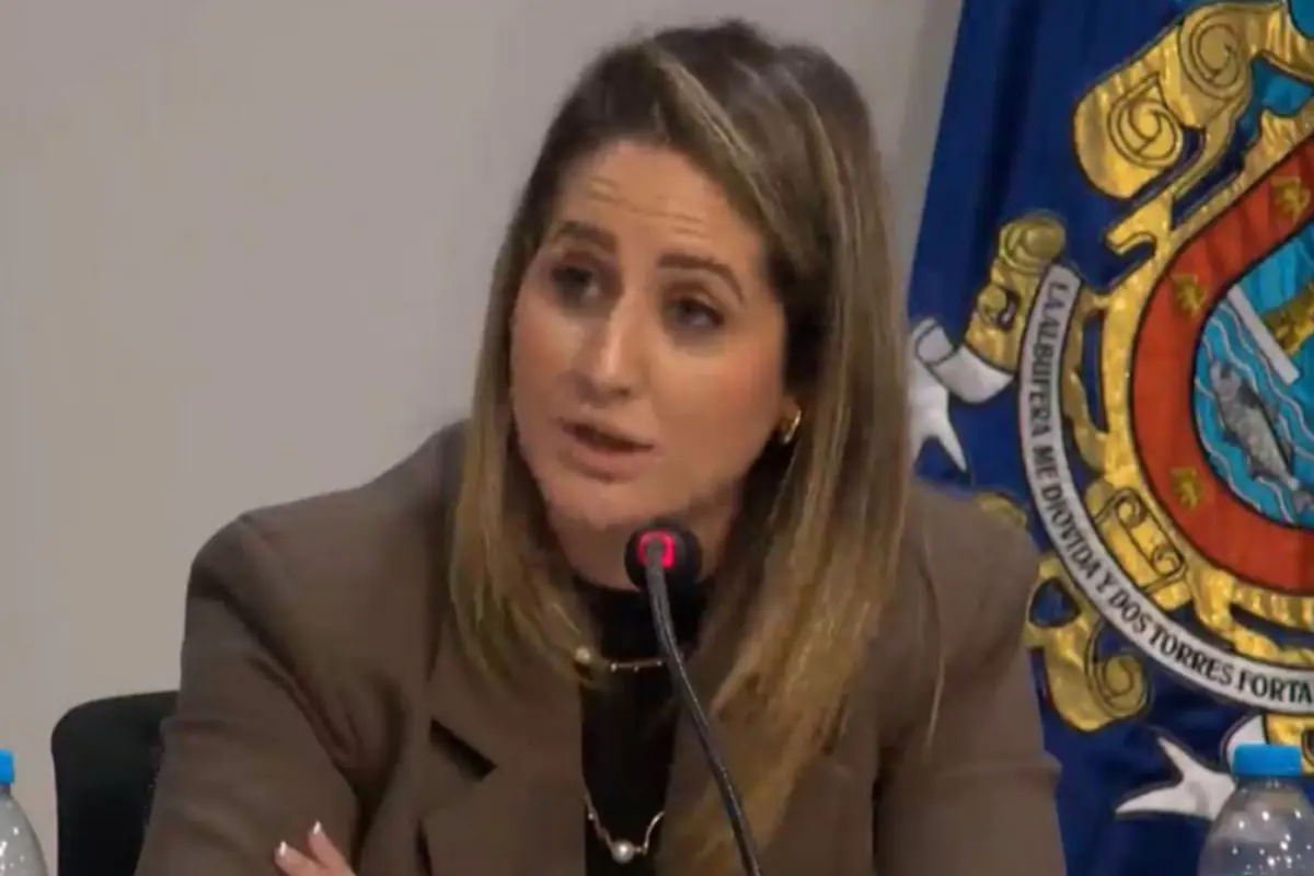 Mujer hablando frente a un micrófono en una mesa de conferencia con un escudo oficial azul y dorado de fondo