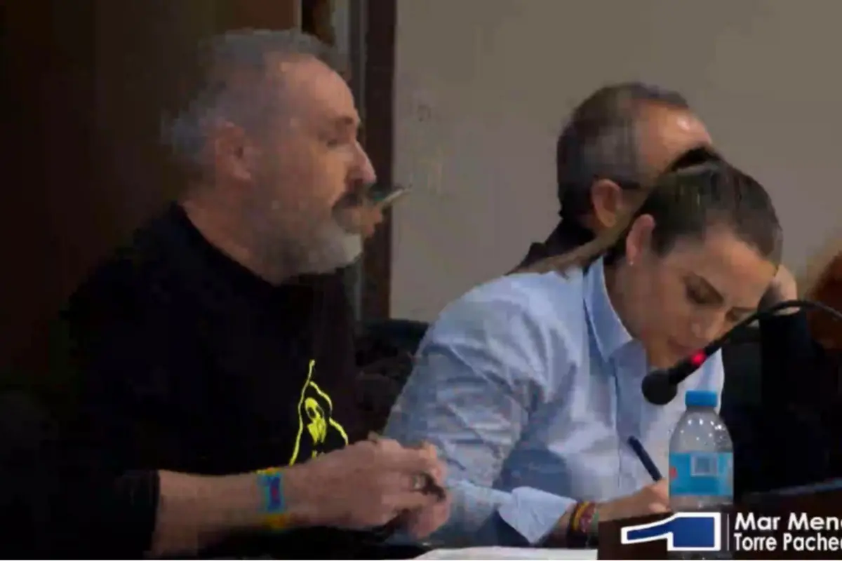 Dos personas sentadas en una mesa durante una reunión o pleno una de ellas hablando frente a un micrófono y la otra tomando notas junto a una botella de agua