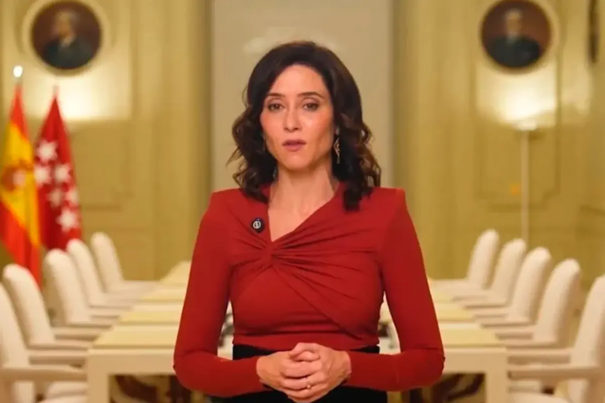 Mujer de pelo castaño con blusa roja hablando de pie en una sala de reuniones oficial con banderas al fondo