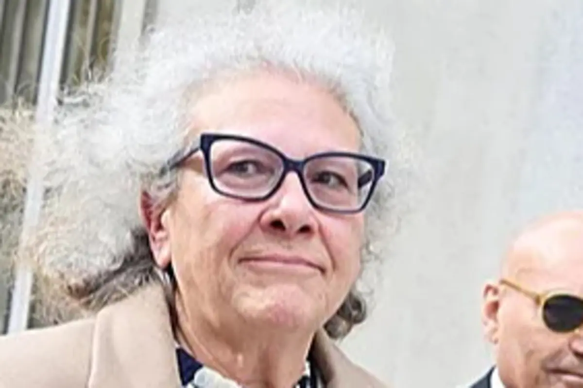 Mujer mayor con gafas oscuras y cabello canoso esponjado mirando a la cámara en un entorno urbano