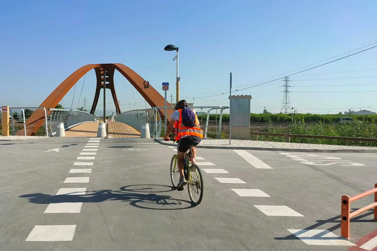 Persona en bicicleta con chaleco reflectante cruzando un puente peatonal y ciclista moderno en un día soleado