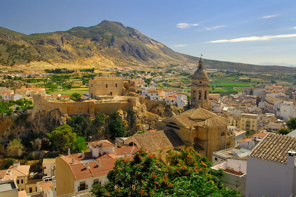Vista panorámica de un pueblo mediterráneo con iglesia de torre alta, muralla antigua sobre un risco y montañas áridas al fondo