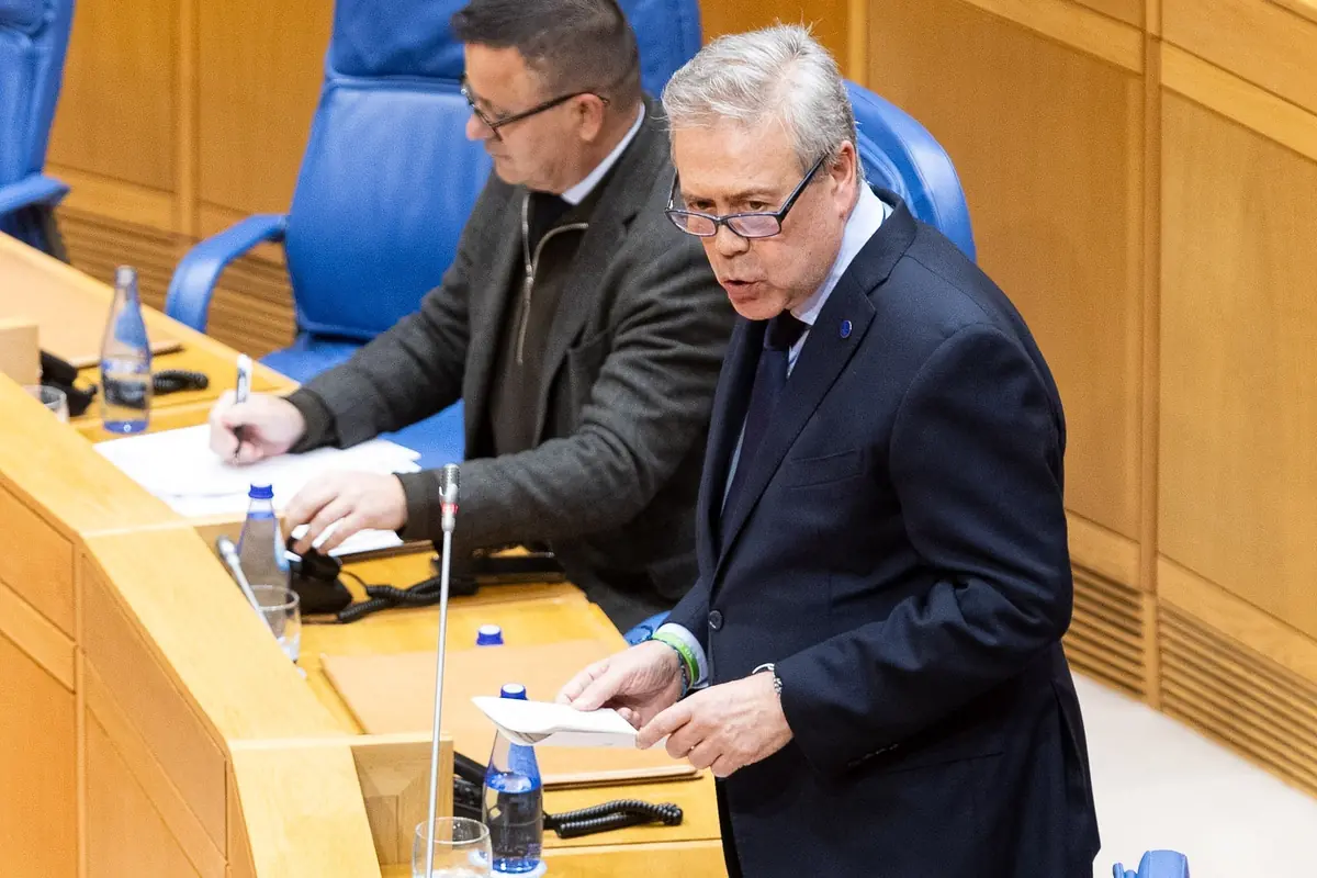 Hombre de cabello canoso hablando mientras sostiene unos papeles en una sesión parlamentaria con otro hombre tomando notas a su lado Hombre de cabello canoso hablando mientras sostiene unos papeles en una sesión parlamentaria con otro hombre tomando notas a su lado