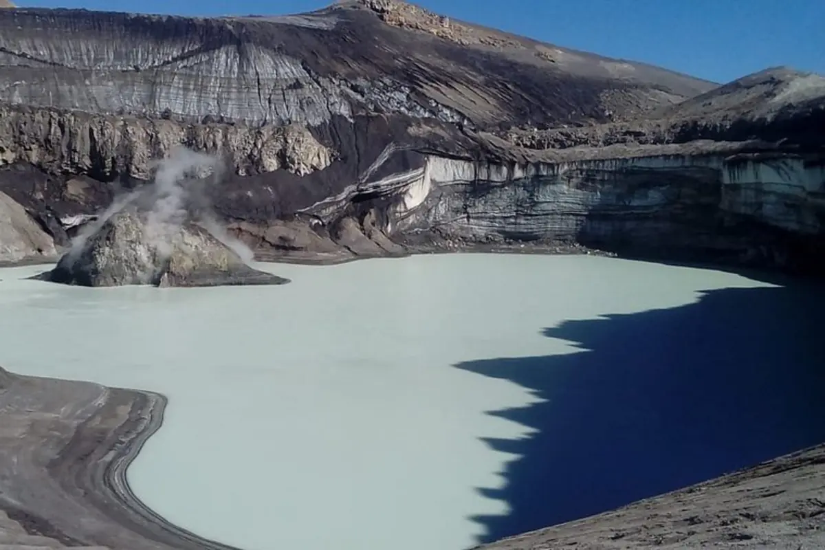 Lago cratérico de aguas claras dentro de un volcán con una isla rocosa humeante y paredes oscuras alrededor