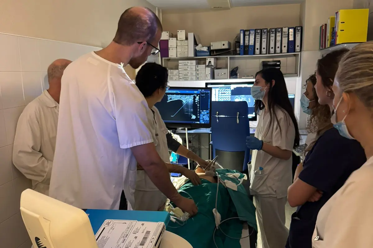 Personal médico realizando una ecografía guiada en un paciente mientras varios profesionales observan la pantalla del monitor en una sala clínica equipada con material sanitario