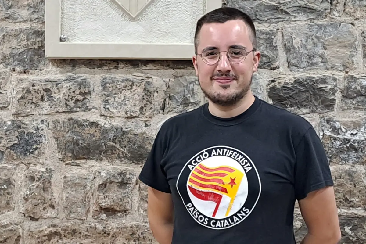 Persona joven con gafas y camiseta negra con un emblema antifascista frente a una pared de piedra