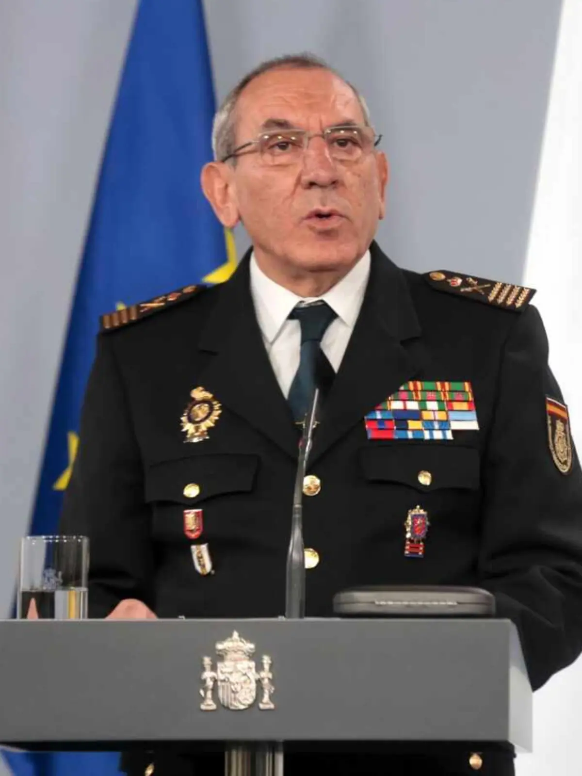 Alto mando policial español con uniforme de gala y condecoraciones hablando en un atril oficial frente a banderas europeas y de España
