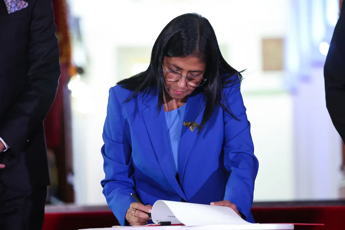 Mujer con gafas y traje azul firmando un documento en un acto oficial