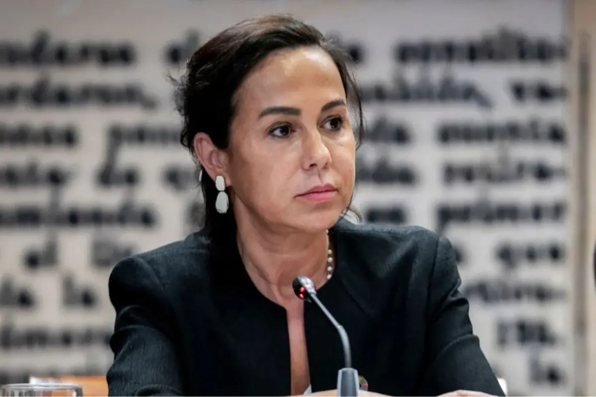 Mujer de mediana edad con saco oscuro y aretes claros sentada frente a un micrófono en una sala de conferencias con texto desenfocado de fondo