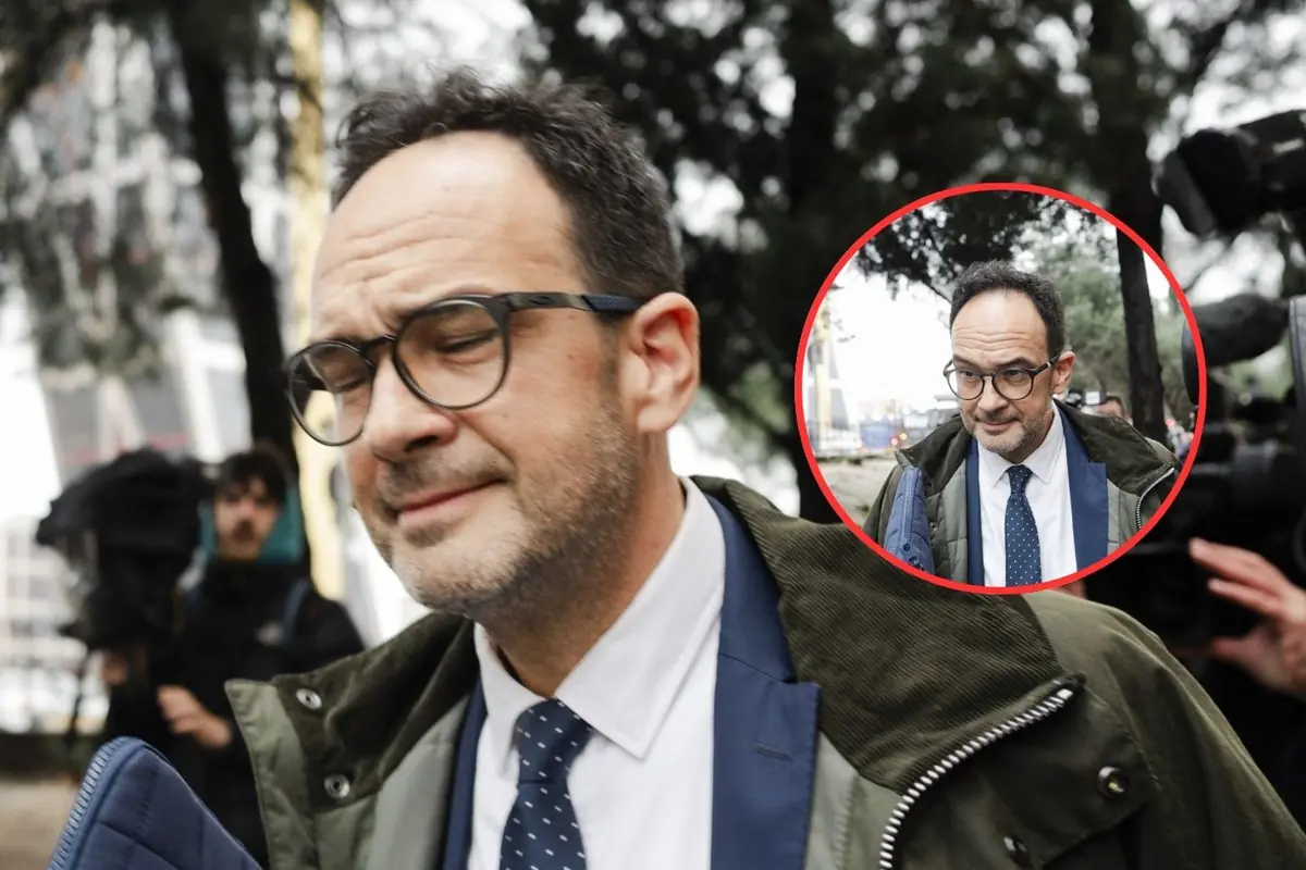 Hombre con gafas y traje caminando al aire libre con gesto de incomodidad mientras es fotografiado por la prensa