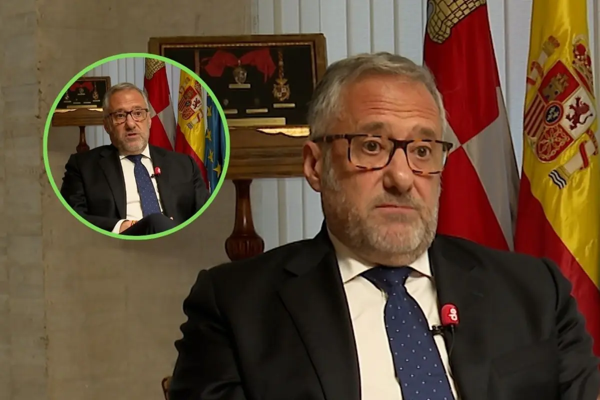 Hombre de mediana edad con gafas y traje oscuro hablando en una entrevista en un despacho oficial con banderas de España y otras banderas autonómicas al fondo y un recuadro circular que muestra otro plano de la misma entrevista