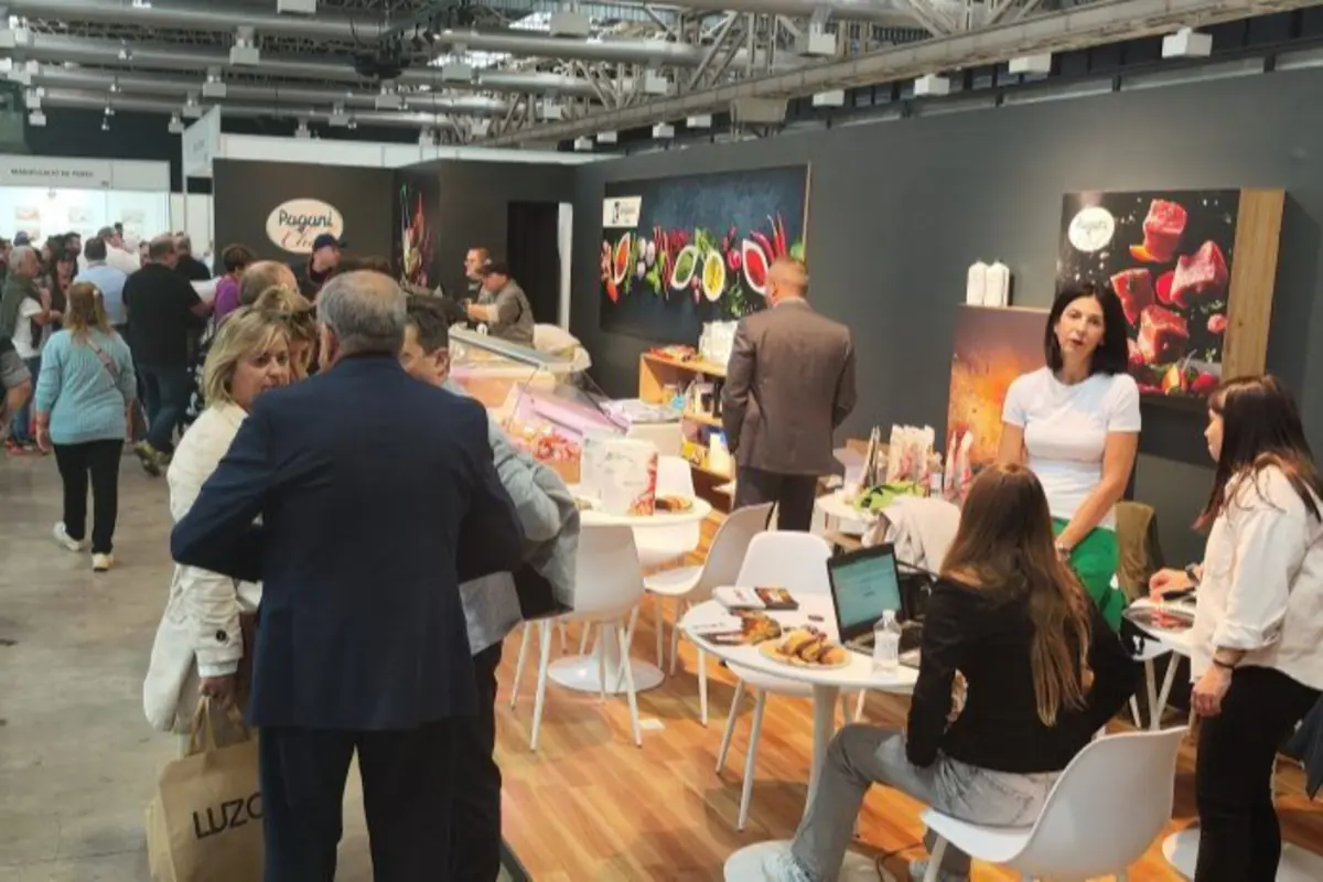 Personas conversando y trabajando en un stand de feria gastronómica con mesas, portátiles y carteles de alimentos de fondo