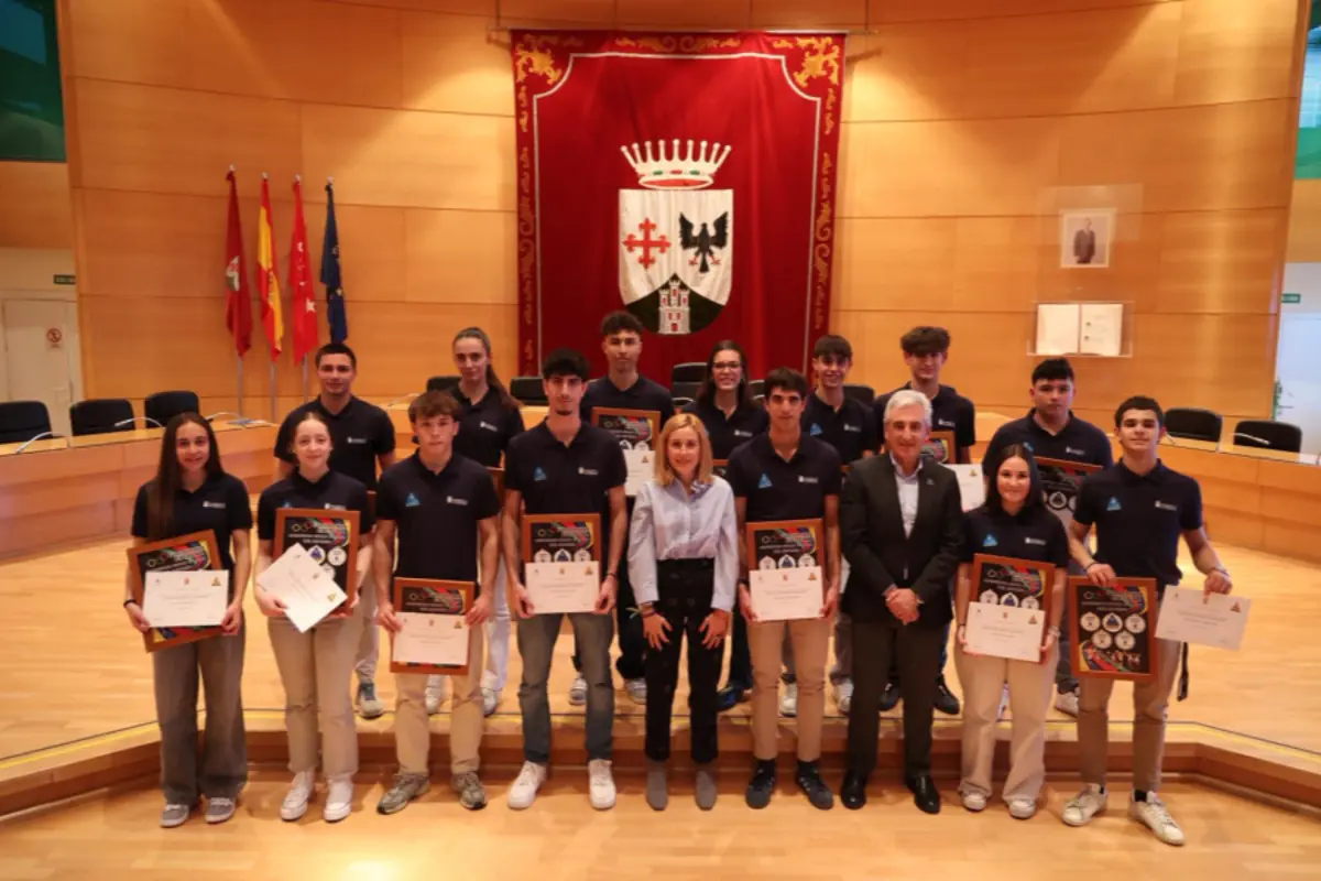 Grupo de jóvenes deportistas y dos adultos posan en un salón de actos oficial sosteniendo diplomas y placas de reconocimiento frente a un gran estandarte rojo con escudo heráldico