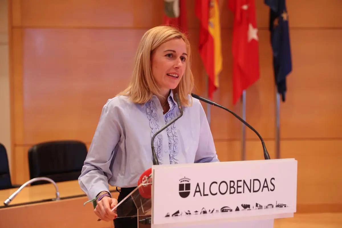 Mujer rubia hablando en un atril con el logotipo y la palabra Alcobendas en una sala institucional con banderas al fondo