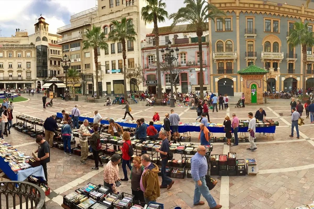 Mercado de libros al aire libre en una plaza urbana con puestos llenos de libros y muchas personas paseando y comprando rodeadas de edificios históricos y palmeras