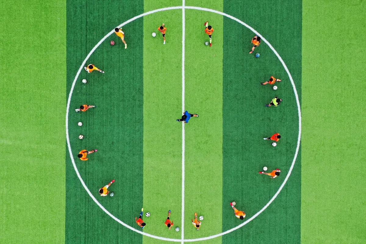 Vista aérea de un grupo de jóvenes futbolistas formando un círculo alrededor del centro del campo mientras entrenan con balones sobre césped verde a rayas Vista aérea de un grupo de jóvenes futbolistas formando un círculo alrededor del centro del campo mientras entrenan con balones sobre césped verde a rayas