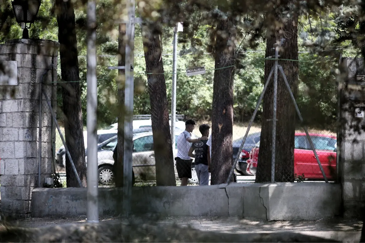 Dos jóvenes conversan junto a una valla entre árboles en una calle con coches aparcados al fondo