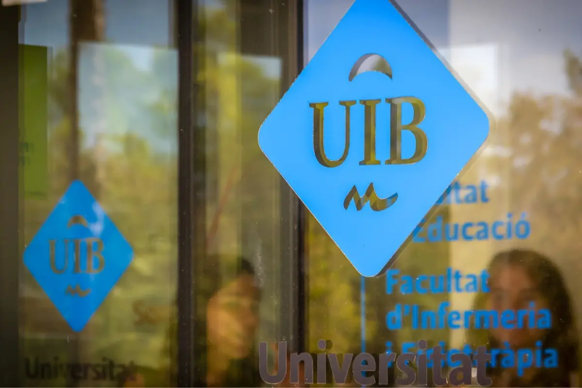 Logotipo azul de la UIB en la puerta de entrada de una facultad universitaria con texto en catalán reflejado en el cristal Logotipo azul de la UIB en la puerta de entrada de una facultad universitaria con texto en catalán reflejado en el cristal