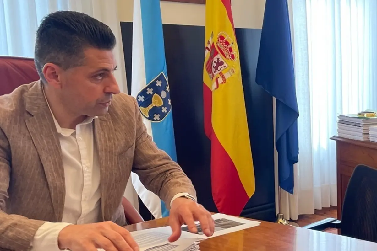 Hombre con chaqueta beige trabajando con documentos en un despacho oficial junto a banderas de Galicia España y la Unión Europea