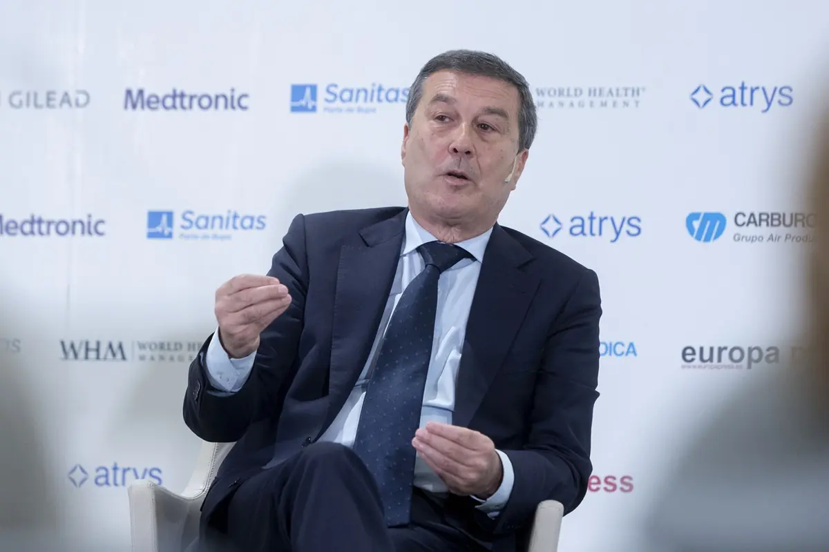 Hombre de mediana edad con traje oscuro hablando gesticulando con las manos durante un evento corporativo con logotipos de empresas de salud en el fondo