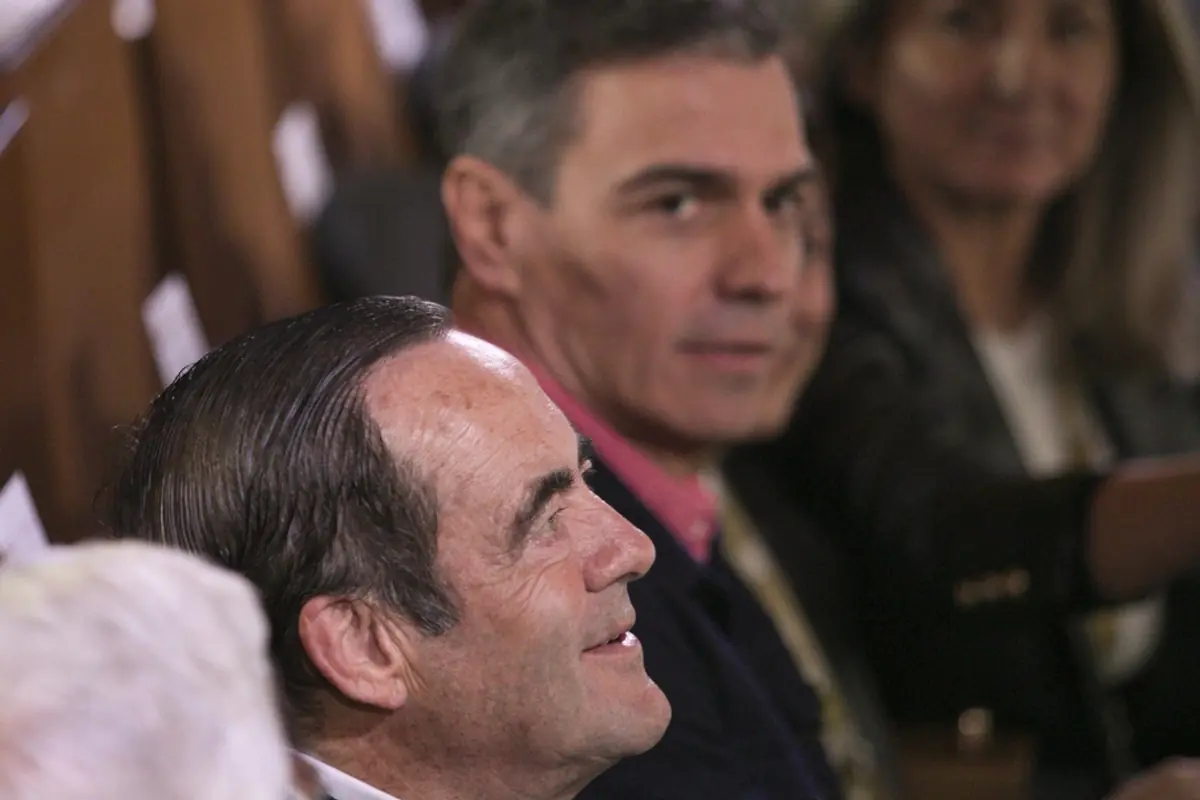 Hombre de perfil sonriendo en primer plano mientras otro hombre al fondo mira hacia la cámara en un ambiente de evento o reunión