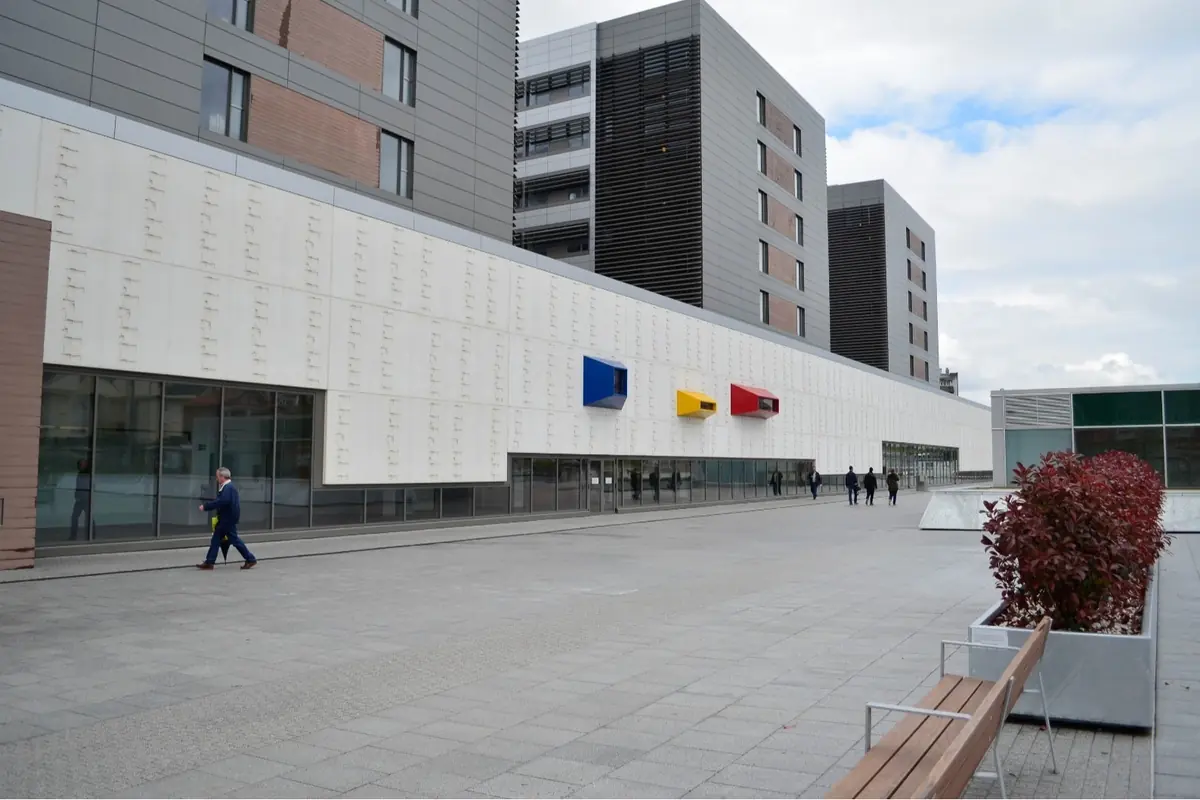 Fachada moderna de un edificio gris y blanco con volúmenes salientes azul amarillo y rojo y una amplia plaza peatonal al frente