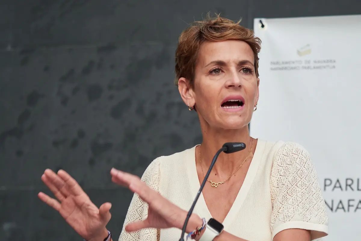 Mujer de mediana edad hablando en un atril con micrófono gesticulando con las manos frente a un fondo institucional