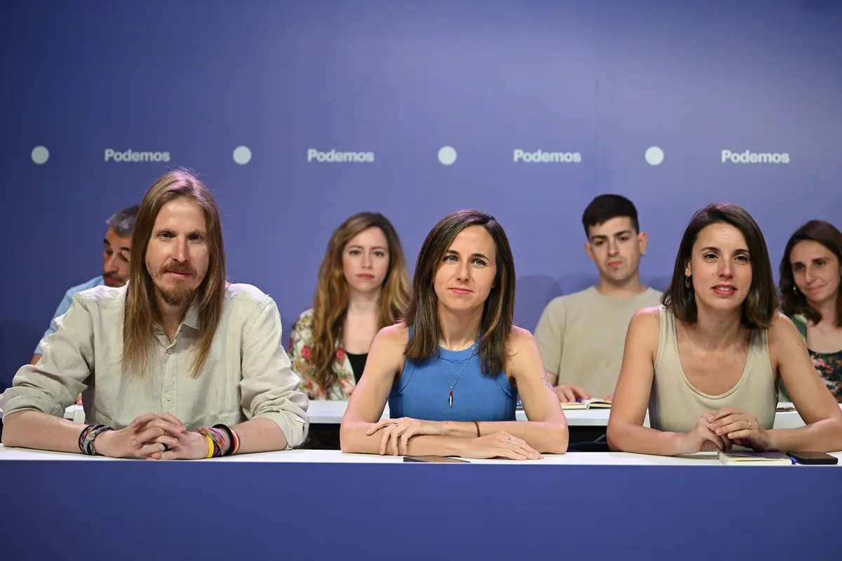 Grupo de personas sentadas en una mesa frente a un fondo morado con el logotipo de Podemos