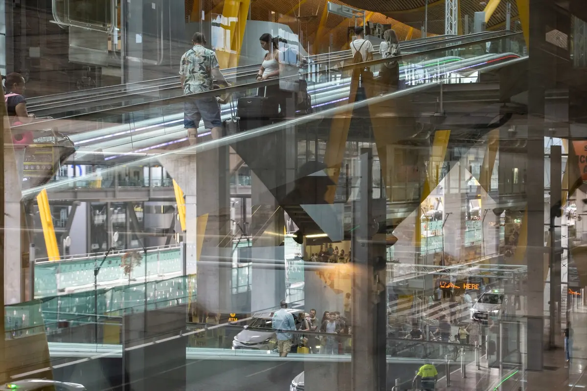 Interior de un aeropuerto moderno con varias pasarelas mecánicas de cristal y personas caminando con maletas entre estructuras metálicas y reflejos en los ventanales