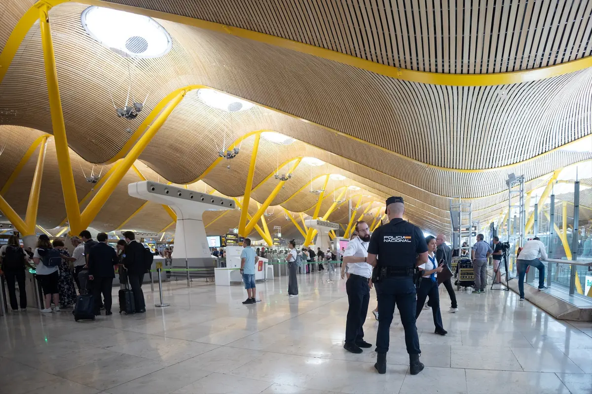 Interior luminoso de un aeropuerto moderno con techo ondulado de madera y columnas amarillas donde pasajeros hacen fila y un agente de policía conversa con varias personas