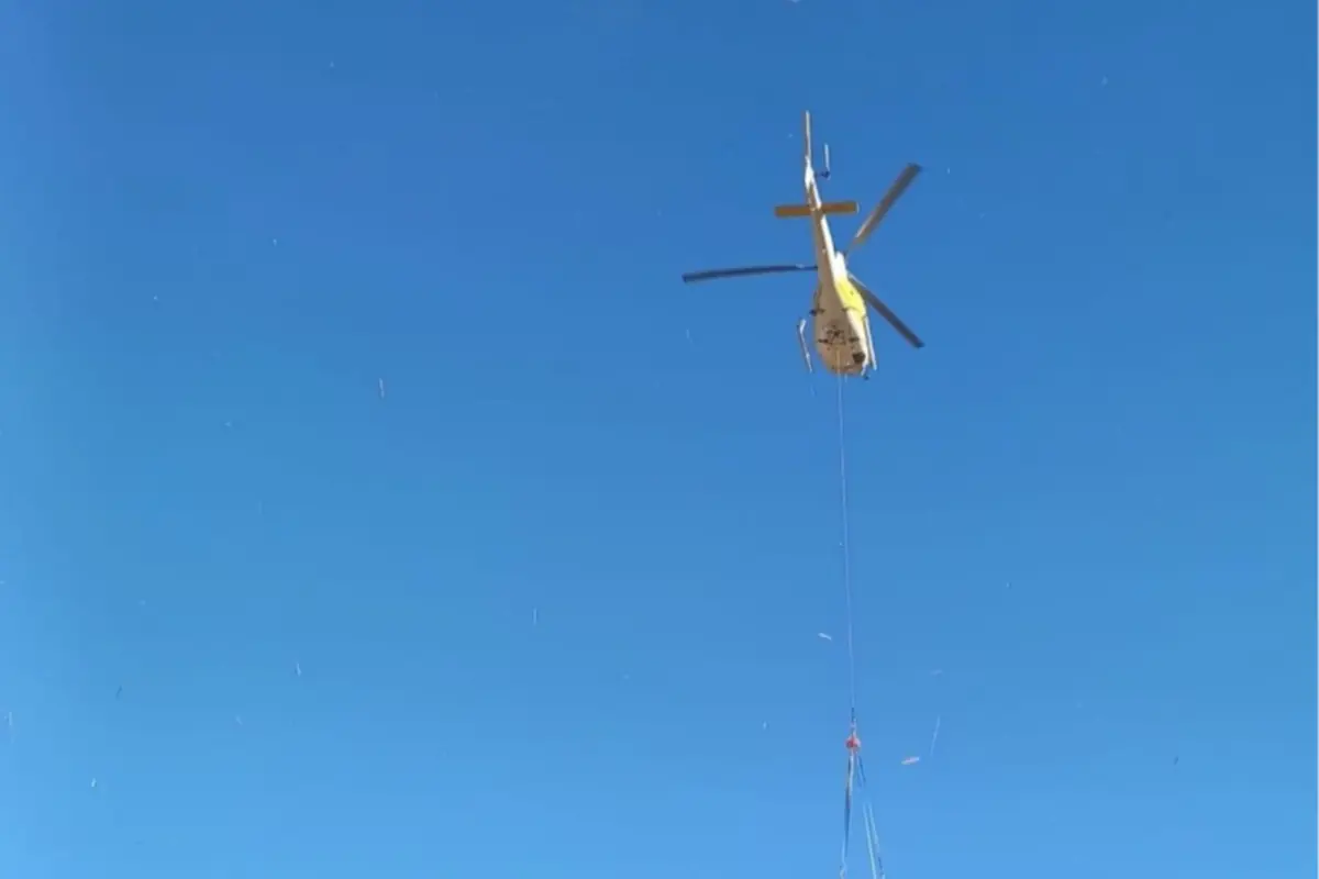 Helicóptero amarillo visto desde abajo transportando una carga colgada con cable en un cielo azul despejado