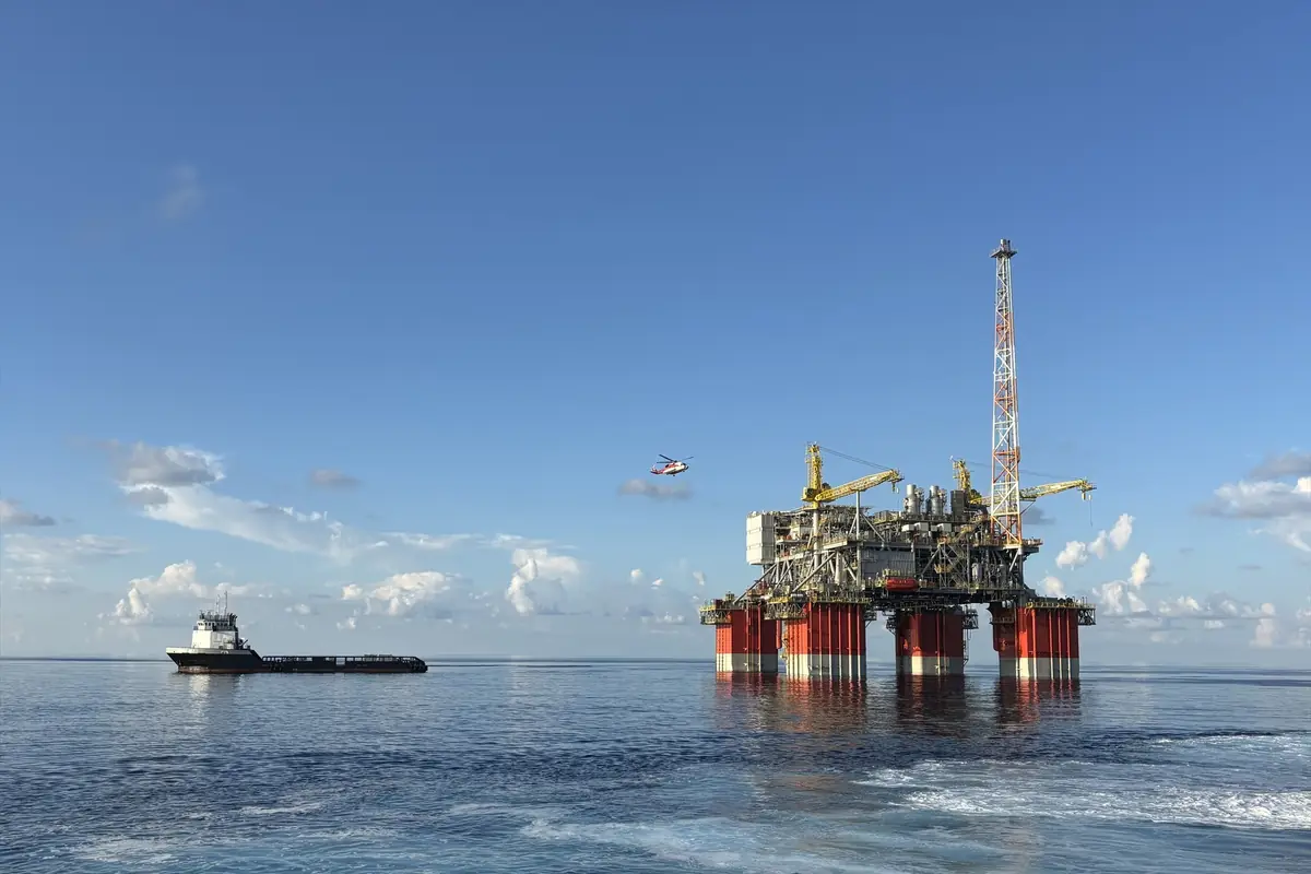 Plataforma petrolera marina con grúas amarillas y base roja en medio del océano junto a un barco de apoyo y un helicóptero volando sobre un cielo despejado
