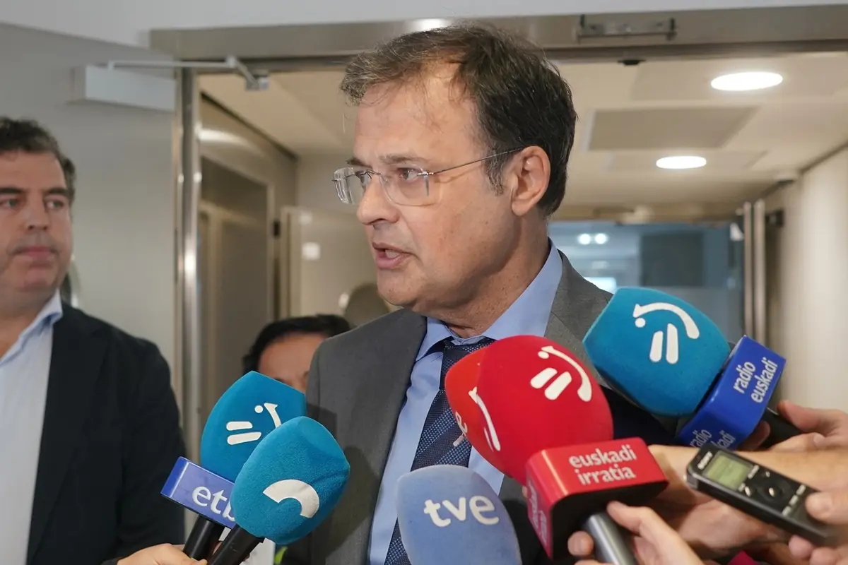 Hombre con traje y gafas atendiendo a varios micrófonos de diferentes medios de comunicación en un pasillo interior