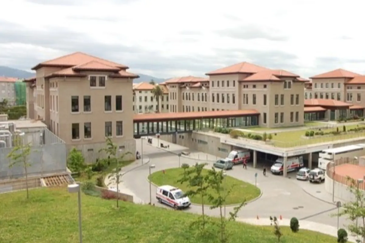 Complejo hospitalario moderno con varios edificios de tejado rojo, zonas ajardinadas y ambulancias circulando por la entrada principal
