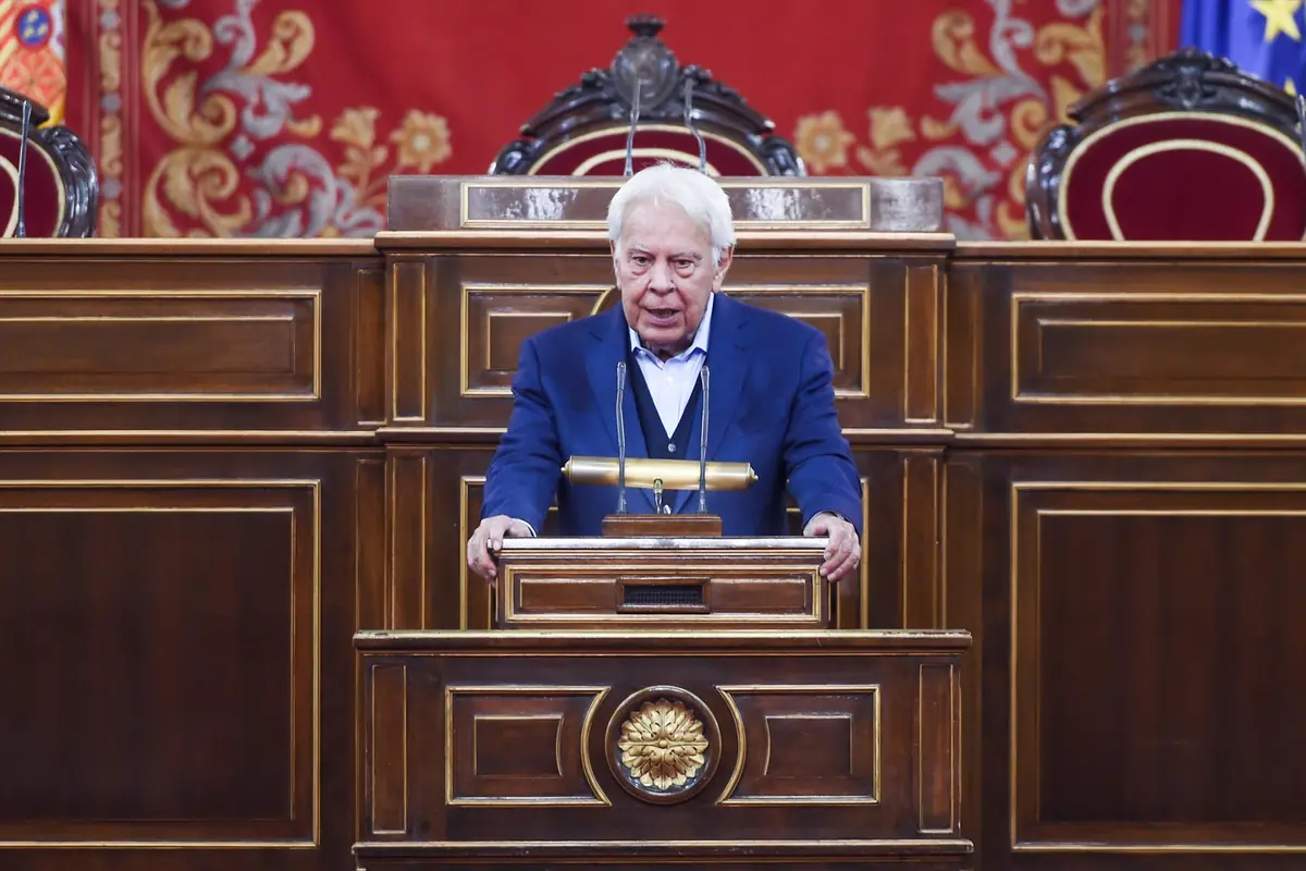 Hombre mayor de cabello canoso hablando en un atril de madera en un salón parlamentario con decoración roja y dorada